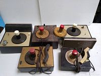 Five vintage RCA turntables arranged on a white surface for viewing, showing various models with buttons and knobs on top, speaker grills, and attached cords