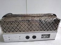 Side view of tube amplifier showing metal casing with geometric ventilation holes and power cable attached.