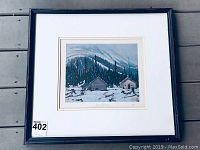 Framed print visible in black frame with white mat showing snow-covered cabins and forested mountain landscape under cloudy sky.