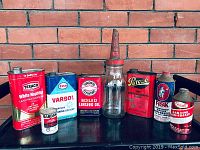 All seven vintage advertising tins and a glass oil bottle arranged in a single row against a brick wall background. The tins feature various brand names and different colors with signage and print design.