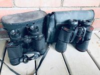 Two pairs of vintage binoculars side-by-side with their respective carrying cases on a wooden surface against a brick wall.