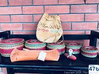 Overall view of deerskin bag, woven baskets, cedar canoe, and bird figurine together on a black chair against brick wall.