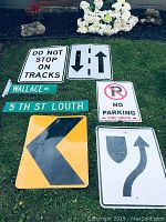 Seven street and traffic signs arranged on grass including directional arrows, street names, parking, and traffic safety signs.