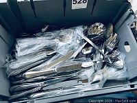 Grey bin containing assorted stainless steel flatware items mostly wrapped in plastic bags, including spoons and knives.