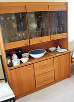 Front view of the teak veneer hutch and buffet with various kitchenware placed on the buffet top and inside hutch shelves behind glass doors.