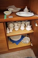 Assorted plates, bowls, lidded sugar bowl, tiered stand rod, cups and saucers displayed on wooden shelves.