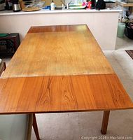 Photo of teak veneer dining table with one hidden leaf extended showing surface wear, scratches, fading, and stains.