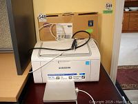 Samsung ML-2165 monochrome laser printer shown with power cables and external device attached, placed on a dark surface with beige surroundings.