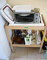 Oster toaster oven on top of wooden table, with various kitchen appliances below including food processor, hand mixer, mini chopper, and baking pan.