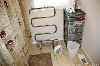 Electric Warmtails towel warmer positioned near window with metallic serpentine shape and reusable base. Shower curtain with floral print partially visible. Adjacent plastic storage bin with five transparent drawers filled with various items. Small white Bellagie fan and white waste bin on floor.