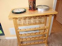 Full view of wooden wine rack side table with top holding two clocks and terracotta wine cooler. Wine glasses hanging below top shelf.