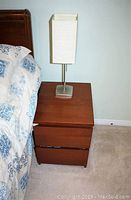 Photo of a wooden night table with two drawers, medium brown finish, with a modern table lamp that has a metal base and a square white fabric shade.