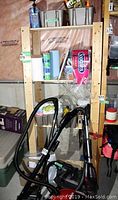 Wood shelving unit with three shelves holding paper towels, cleaning supplies, and small containers, light wood color, minor wear visible.