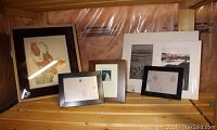 Multiple framed artworks including a coloured pencil drawing with glass glare, two larger black and white prints, and smaller photo frames on wooden shelf