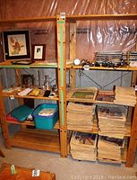 Photo showing two wooden shelving units with items stored on shelves, including framed art, glass bottles, and stacked papers.