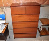 Front view of tallboy dresser showing six drawers, wood-tone finish, setting in basement area.