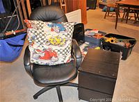 Black vinyl office chair with oversized colorful pillow featuring race cars and checkered design on the seat, black MDF side table with two drawers beside chair on concrete floor