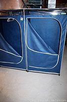 Side view of two navy blue portable closets with white trim, zippered fabric doors partially open revealing interior space. Shown on concrete floor.