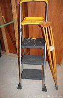 Full view of the Cosco folding step ladder with yellow painters tray and a pair of wooden crutches leaning against the ladder.