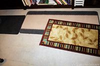 Three polypropylene area rugs displayed on floor: beige/grey striped rug with zigzag edges, Daniella runner with leaf design and multicolor border, solid dark grey rubber backed rug.