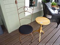 Metal chair with arrow design and black cushion seat next to wooden side table with tripod legs on porch floor.
