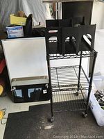 Photo showing whiteboard leaning against a wall with black metal cart containing black file folders on top and bottom shelves, and tabletop TV mount on shelf