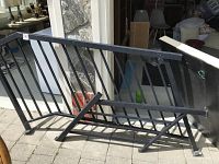 Larger black metal railing measuring about 72x39 inches, angled mounting ends, vertical bars, resting outside on pavement near a stone wall and glass door.