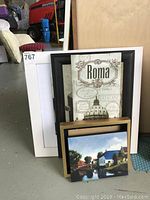Three framed prints stacked together, largest with 'Roma' text and architectural illustration in black frame, medium colorful house painting with light wooden frame, smallest print with white frame.