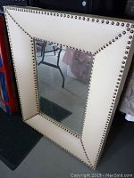 Frontal view of the rectangular bone-colored framed mirror showing brass tacks along the edges and reflective glass in the center.