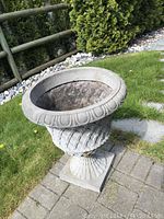 Full view of the outdoor garden urn placed on a stone-paver area with grass and shrubs in background.