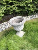 Front angled view of the garden urn placed on grass, showing the urn's carved decorative patterns and pedestal base.