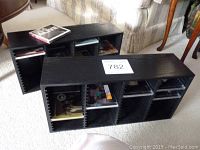 Two black wooden CD racks placed on carpet with various CDs inside the slots. CDs are visibly new in cellophane wrappings.