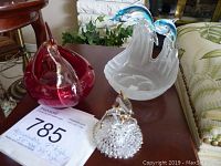 Photo showing three items on a wooden table near a fireplace: a frosted white glass dolphin candle holder with blue dolphins, a red art glass bird figurine, and a clear crystal dolphin figurine with textured base.