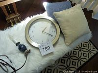 Clock with white face and brushed metal frame, two decorative pillows, electric candle lamp on a faux fur surface.