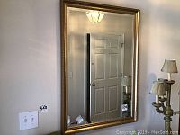 Full view of wall mirror hung on wall showing gold frame and reflection of door and lamp.
