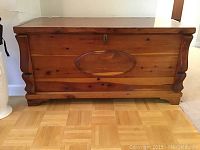 Front view of wooden cedar chest showing central oval decorative panel, wear and nicks on wood surface.