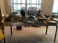 Multiple silver plated and sterling silver items laid out on a table, including trays, pitchers, bowls, plates, and candle holders.