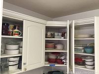 First cabinet with multiple shelves holding various CorningWare dishes, coffee mugs, and glass bowls.