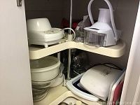 Photo of a kitchen corner shelf inside a cabinet showing a white mini food processor or chopper attachment, stack of clear glass bowls, white plastic cutting boards, and parts of waffle irons.