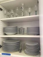 Shelves inside kitchen cabinet filled with glass stemware on top shelf, bowls and plates on middle shelf, and dinner plates and saucers on bottom shelf.