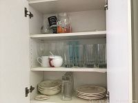 One open cabinet shelf showing multiple clear drinking glasses, blue-tinted glasses, white mugs, all on a white cabinet shelf with more dishes below.