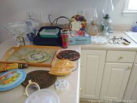 Overview of kitchen countertop showing various items including oil lamps, cake plates, wooden tray, trivets, utensils, and small kitchenware items.