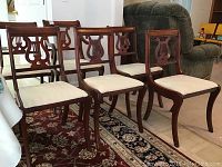 Five wooden dining chairs; four without arms, one armchair with carved lyre motif backs and cream upholstered seats showing some wear and stains.