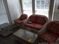 Off white plastic wicker love seat, chair, and rocker with red floral cushions arranged near windows. Includes rectangular coffee table with glass top.