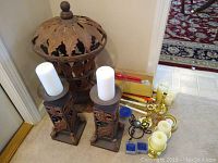 Pair of tall ornate metal candle holders with white pillar candles, large leaf motif metal holder behind, various brass and small holders on floor