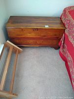Cedar chest closed with small wooden rack beside it on carpeted floor next to a bed with red bedding.