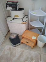Photo showing the entire lot: white woven hamper, Therabath Pro device in box on top of hamper, mechanical scale, Eddie Bauer wooden step stool, white 3-tier corner shelf, bath rug on floor