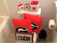 Three red UGA throw pillows stacked with a UGA logo, three-piece ceramic bathroom set with UGA branding on top of toilet tank