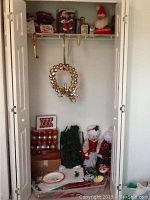 Closet shelf and floor showing various Christmas decorations including a red and gold wreath with bells and reindeer, boxes of red ornaments, plush Santa, snowman figurines, and artificial greenery.