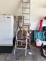 Photo showing full view of extra tall aluminum extension ladder, two wooden ladders, metal step stool, and some plastic items on the right.